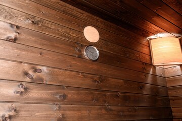 warm sauna interior features wood-paneled walls, a thermometer, an air vent, and a square lamp. The...