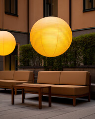 Outdoor seating area with modern brown couches, wooden table, and large glowing yellow spherical lanterns against a backdrop of greenery and building walls.