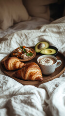 A wooden tray holds fresh croissants, greens, and a cup of coffee, all resting on a bed with soft, white linens. Perfect for a relaxed morning meal at home