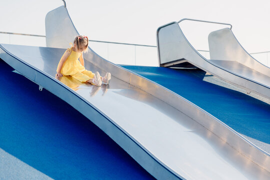 Little girl in yellow dress on modern playground slide, enjoying sunny summer day outdoors. - Powered by Adobe