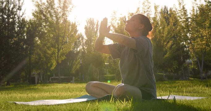 Woman doing yoga outdoors at sunrise with her hands together. Woman sitting on a mat in a green park breathing deeply. Outdoor yoga unites body and mind. Outdoor yoga inspires harmony.