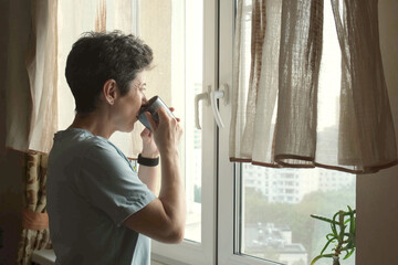 Middle-aged woman standing alone at home, sipping coffee and staring out the window, symbolizing solitude and quiet introspection in urban life