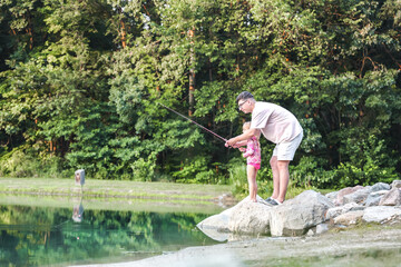 Father and daughter fishing together by a serene lake. A heartwarming moment of bonding in nature, sitting on rocks near the water. Family outdoor activity in summer.