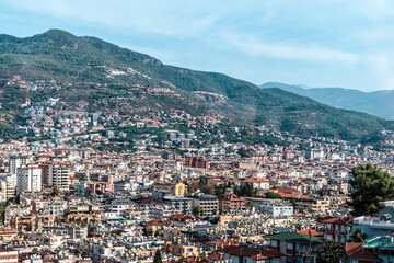 Panoramic view of Alanya, Turkey, with the city's buildings stretching across the landscape and the Alanya sign visible on the hillside. The backdrop includes green mountains under a clear sky