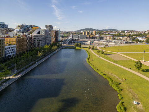 Aerial view of Barcode buildings reflected in the shimmering waters of the fjord, juxtaposed against the green parkland and distant hills, Oslo, Norway.