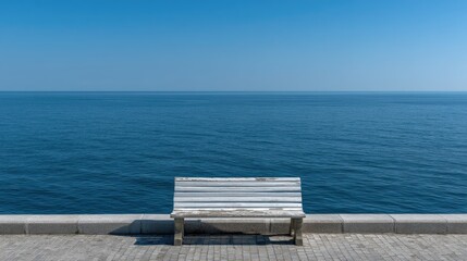 Empty white wooden bench on a waterfront walkway, facing a vast expanse of blue ocean under a clear sky