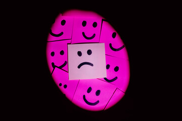 Sad face surrounded by happy faces drawn on colored paper with black marker with a uniform solid background