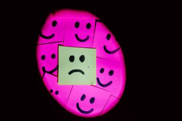 Sad face surrounded by happy faces drawn on colored paper with black marker with a uniform solid background