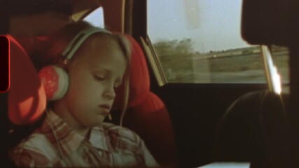 Filmed on super 8 film blonde young girl peacefully resting in car seat, listening to music through headphones during family road trip, countryside landscape blurring outside window