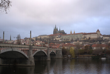View of Prague Castle from the Vltava River with a cloudy sky and bridges in the foreground during late afternoon