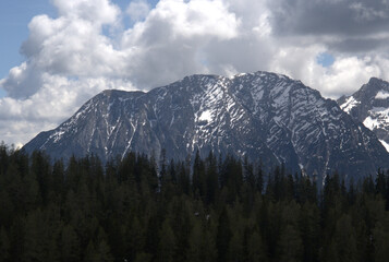 Obraz premium Snow-capped mountain peaks rise above dense evergreen forests under a cloudy sky in the afternoon light