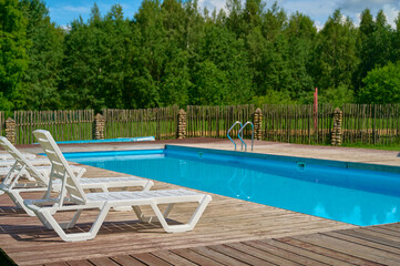 Relaxing outdoor swimming pool with wooden deck, surrounded by lounge chairs and green landscape with dense trees, under clear blue sky during daytime