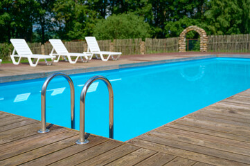 Inviting outdoor swimming pool with wooden deck and modern stainless steel ladder, surrounded by white lounge chairs and lush green trees, sunny daytime atmosphere