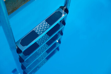 Close-up of modern stainless steel pool ladder descending into clear blue water, with the reflection of steps visible on the water surface during bright daylight