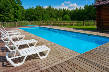 Relaxing outdoor swimming pool with wooden deck, stainless steel ladder, and white lounge chairs, set amidst lush green landscape and a clear blue sky during daytime