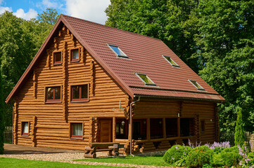 Rustic wooden log cabin with steep gable roof, surrounded by green lawn and blooming flowers,...