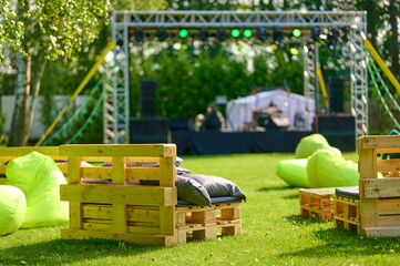 Relaxed outdoor event space on lush green lawn with wooden pallet lounge chairs, bright green bean bags, and a stage with lighting, trees, and clear sky atmosphere