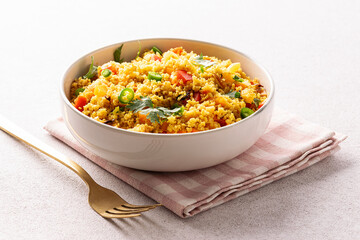 Healthy homemade vegan couscous salad with cilantro, colorful bell peppers pieces, green chilli pepper and spices in ceramic bowl on light stone table close up.