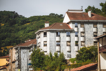 Modern residential buildings with light stone facades and red tiled roofs in the town of Tineo, Spain, surrounded by lush green hills under clear daylight in a peaceful rural setting