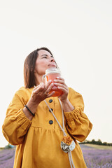 Woman in yellow dress holding lavender flower and jar of honey while looking up in purple field