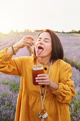 Smiling woman in lavender field enjoying sweet honey from lavender flower held above her tongue