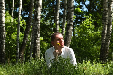 Young man with yellow dandelion in hand lies in the grass in a birch forest.