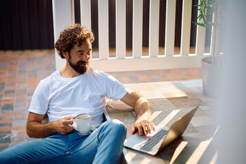 Smiling man with cup of coffee surfing the net on laptop on porch.
