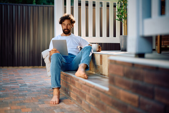 Mid adult man drinking coffee and surfing the net on touchpad while relaxing on porch.