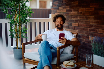 Happy man using cell phone while enjoying in morning coffee on porch.