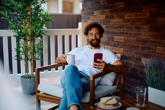 Happy man using cell phone while enjoying in morning coffee on porch. - Powered by Adobe