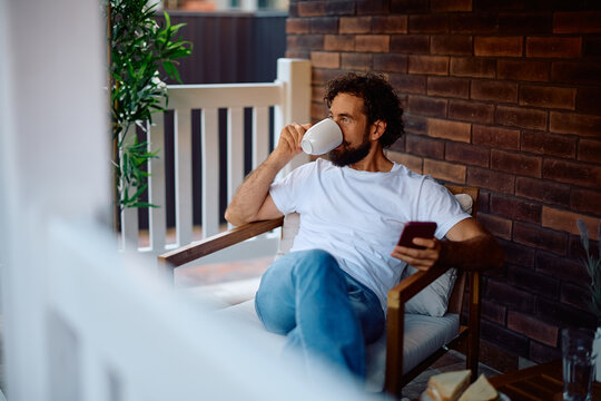 Mid adult man drinking coffee while using smartphone and relaxing in armchair on patio.