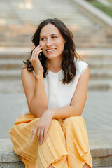 A young woman is enjoying a delightful phone call while sitting outdoors, smiling and feeling...