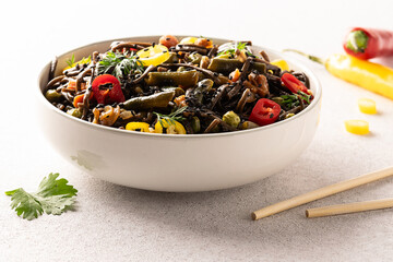 Soba noodles with teriyaki sauce, vegetables and herbs in white ceramic bowl on light stone background close up. Tasty hot Asian food.
