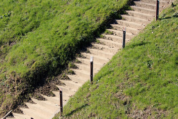 A bare concrete stairway with rusted metal posts ascends a green, grassy embankment, providing functional access up a steep slope in a landscaped outdoor setting