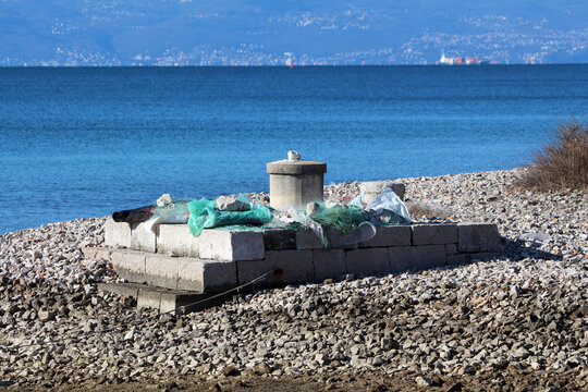 A square base made of concrete blocks and topped with plastic netting and debris lies abandoned on a stony, coastal shoreline, with blue sea and distant city visible across the water