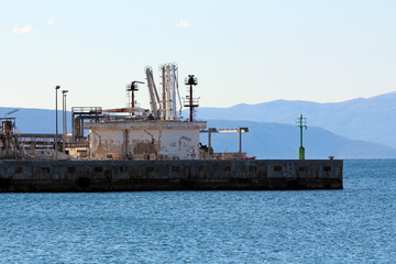 Fototapeta premium An aging dockside marine terminal features rusted equipment and industrial structures set along blue water, framed by distant mountains under a pale sky