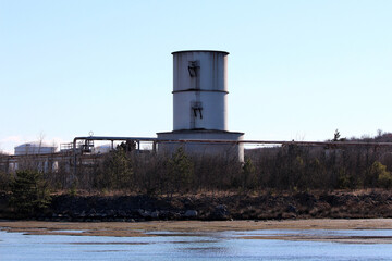 Obraz premium A tall, metallic distillation column with visible ladders dominates the shoreline, surrounded by piping and low vegetation at a coastal refining or chemical facility