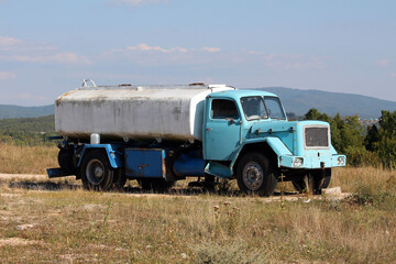 A vintage blue water tanker truck with a large white tank rests on uneven dry grass, its faded paint, rusty details, and missing rear wheels revealing years of service in a rural landscape