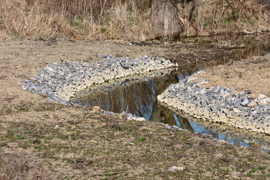 Shallow stream banks are reinforced with layers of angular riprap stones to prevent erosion, with calm water reflecting surrounding dried grass and trees