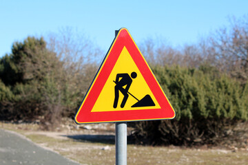 A bright triangular road sign with a black construction worker symbol warns drivers of upcoming roadwork, standing out against green roadside shrubs and a blue sky
