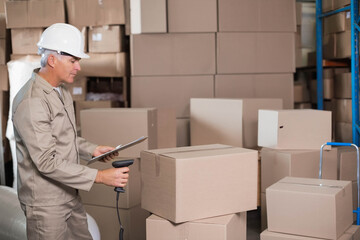 Senior man wearing coveralls scanning boxes at warehouse with barcode scanner and clipboard