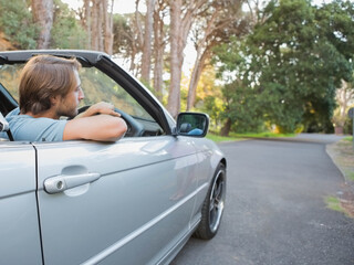 Man resting forearms on door of silver convertible with top down gazing pine-lined road, copy space