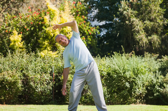 Senior man in sportswear stretching sideways on manicured lawn in garden under bright sunlight