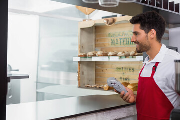 Male cashier wearing red apron offering card reader at bakery counter with bread shelf, copy space