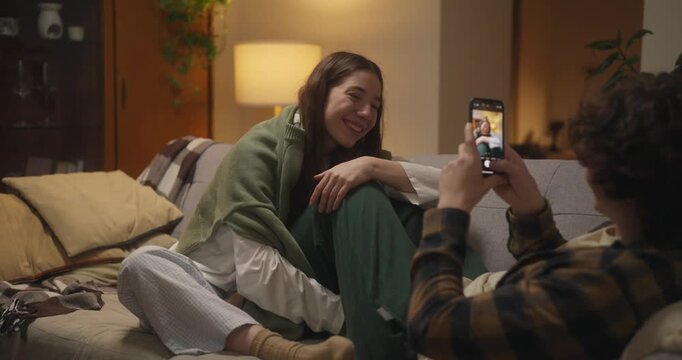 Over the shoulder guy with curly hair takes a photo of his shy girlfriend who poses and covers the phone with her hand during a cozy evening on the couch at home