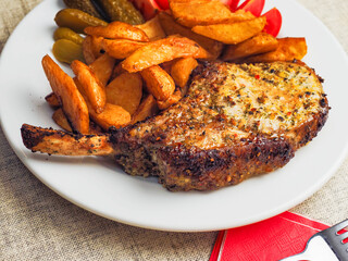 A plate of food with a premium pork chop in the bone, wedges potatoes, and tomatoes. The plate is set on a table with a fork and a knife. Fine tasty dinner with high quality meat.