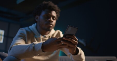 African American man scrolling through social media on smartphone while relaxing on a couch, cozy...
