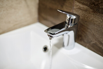 Modern chrome faucet with flowing water in a pristine white sink, surrounded by textured stone wall, showcasing contemporary bathroom design and functionality