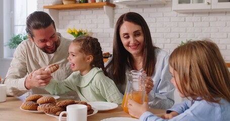 Happy family having breakfast together at kitchen, eating cookies and drinking orange juice, having pleasant talk, wishing each other good day. Caring mother, father and their children having meal - Powered by Adobe