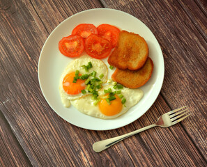 Fried eggs with green onions, fried tomatoes and bread croutons on a wooden table, a fork nearby.